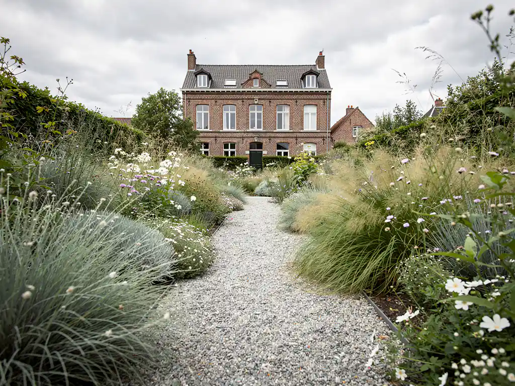 Naturalistische siertuin in volle bloei met siergras en vaste planten langs grindpad naar klassieke bakstenen herenwoning.